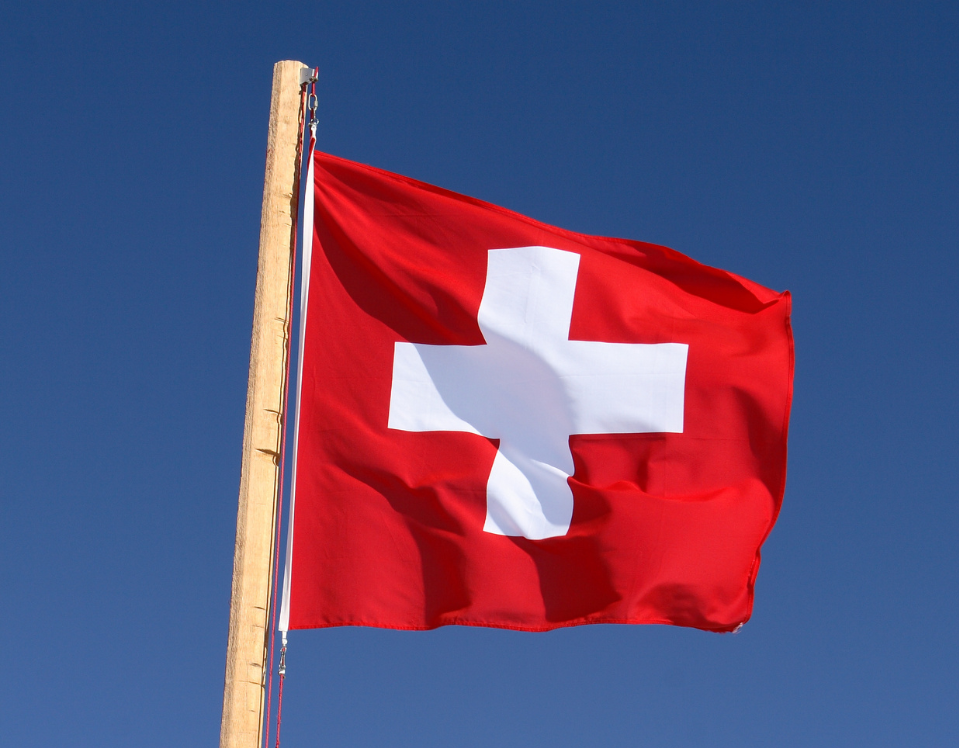 Swiss national flag in front of blue skies on wooden pole.